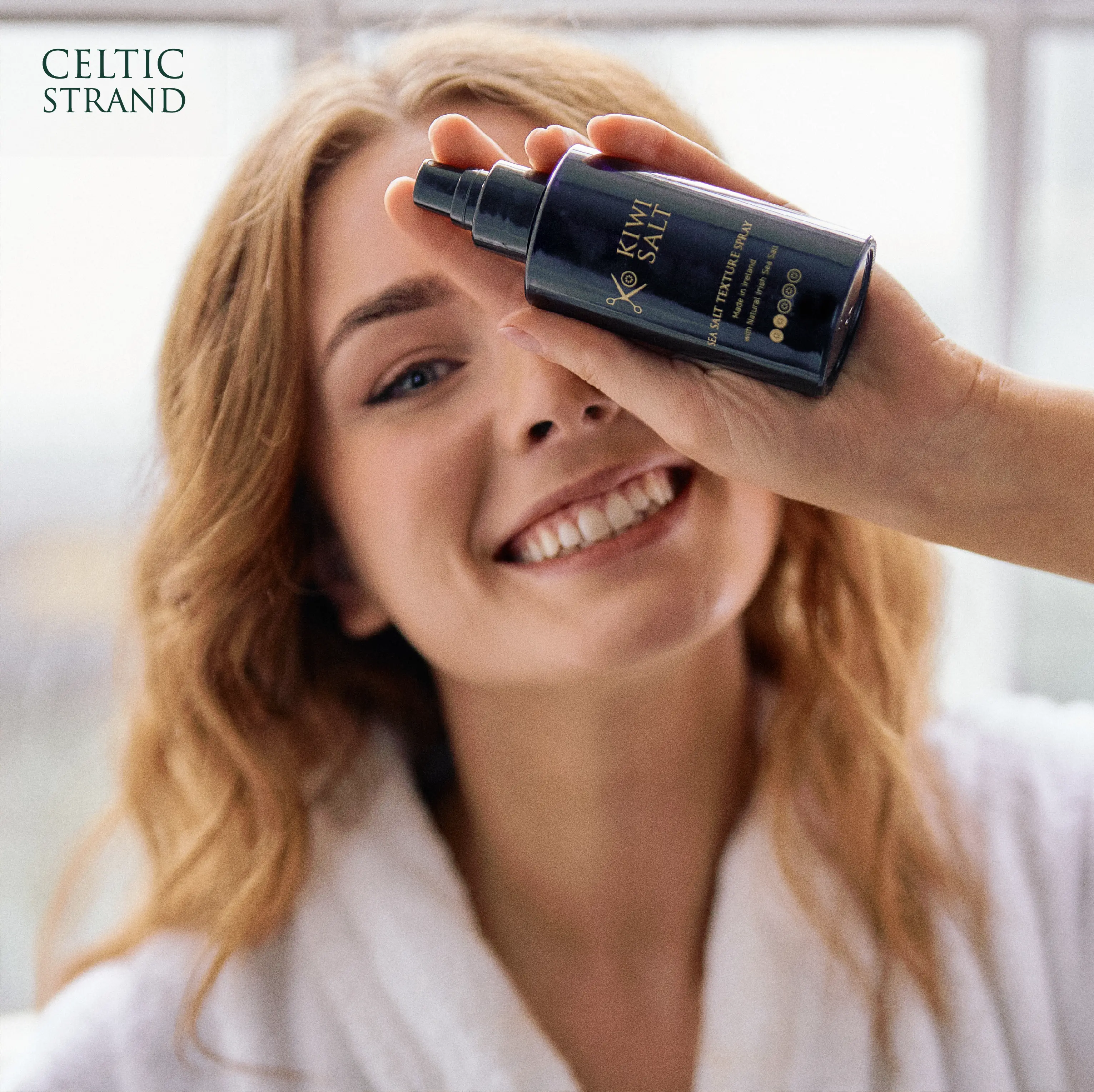 Smiling woman in a white robe with wavy hair holding a KiwiSalt Sea Salt Texture Spray bottle to her face.
